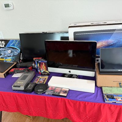 Collection of electronics, media, and tools displayed on a table, including an Apple iMac with original box, ASUS portable monitors,...