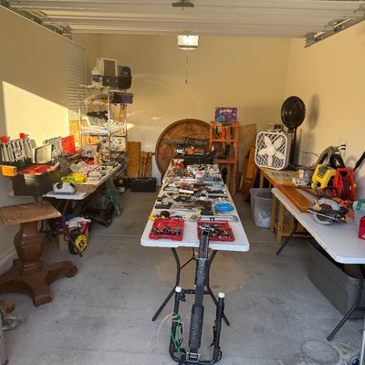 Garage workshop filled with various hand tools, power tools, step ladders, storage units, and fans arranged on folding tables.