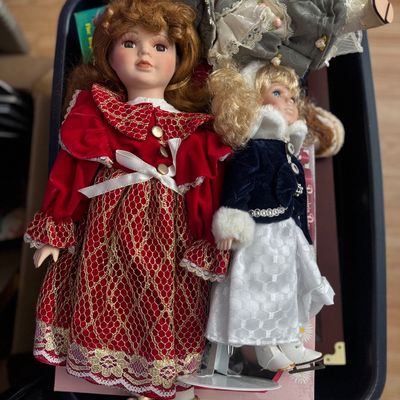 Collection of vintage porcelain dolls in elaborate winter and holiday dresses, displayed together in a storage bin.