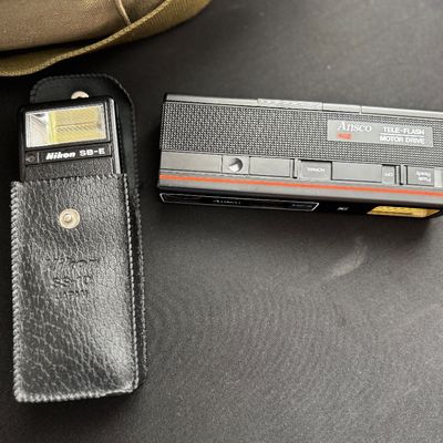 Sale Photo Thumbnail #6: Vintage Nikon SB-E speedlight flash unit in its black leather SS-10 case, shown alongside an Ansco Tele-Flash Motor Drive 412 camera.
