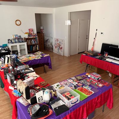 Estate sale room setup featuring tables draped in purple and red cloths, displaying various merchandise including electronics, craft...