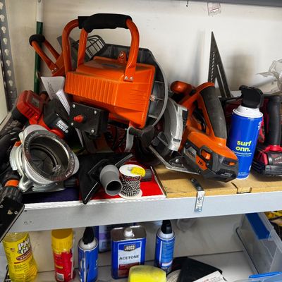 Assorted power tools and workshop supplies, including a Ridgid 18V circular saw, an orange portable work light, and various cleaning...