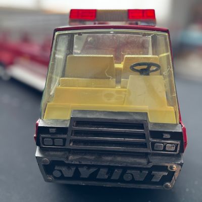 Sale Photo Thumbnail #10: Vintage Nylint pressed steel fire truck toy, featuring a yellow plastic interior, black steering wheel, and red emergency light bar.
