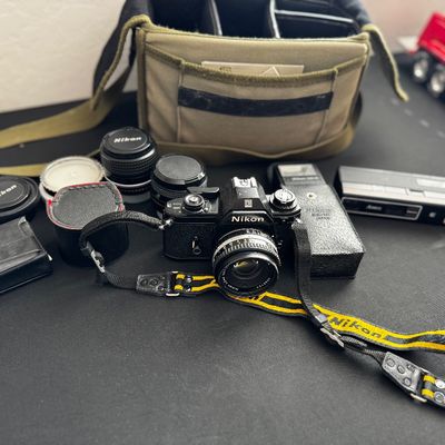 Sale Photo Thumbnail #7: Vintage Nikon EM 35mm SLR camera with a yellow and black Nikon strap, multiple lenses, accessories, and a canvas camera bag.