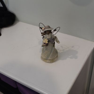 Sale Photo Thumbnail #7: Small decorative angel figurine featuring a woman in a hat holding flowers, with wire wings, displayed on a white shelf.