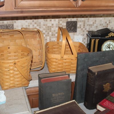 Collection of vintage Longaberger woven baskets, antique books including a Holy Bible, and a black mantel clock with gold-tone columns.