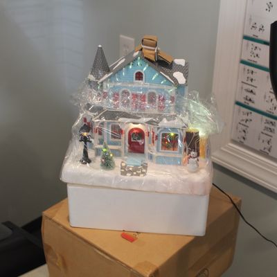 Light-up Christmas village house wrapped in protective plastic, featuring a blue Victorian-style home with a snowman and decorated tree...