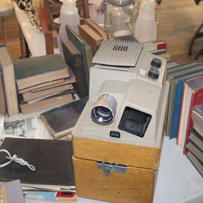 Vintage Wollensak slide projector sitting on its textured carrying case, surrounded by stacks of old hardcover books on a table.