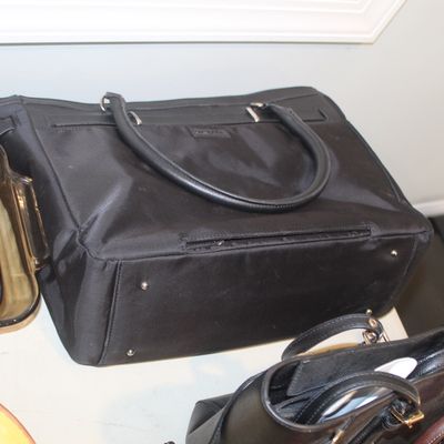 Black nylon tote bag with leather handles and metal feet, shown alongside other handbags and amber glassware.