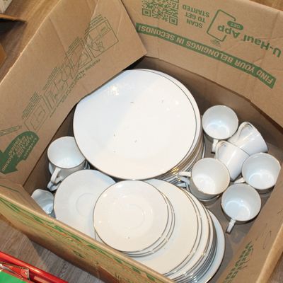 Sale Photo Thumbnail #20: A collection of white ceramic dinnerware with silver rims, including plates, saucers, and teacups, packed inside a cardboard moving box.
