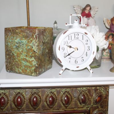 Shabby chic white metal alarm clock with distressed finish, paired with a square textured ceramic planter and various decorative figurines.