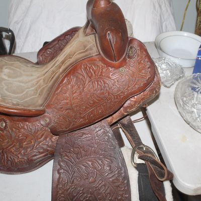 Vintage leather western saddle featuring intricate oak leaf and acorn hand-tooling, displayed on a table alongside various household items.