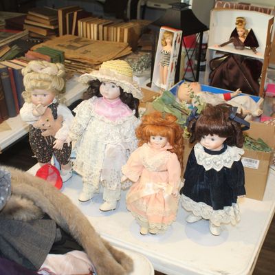 Collection of vintage porcelain dolls in lace dresses, alongside boxed Barbie dolls and stacks of vintage books on a display table.