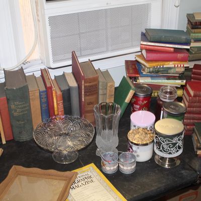 Assortment of vintage hardcover books, glass vases, candles, and a National Geographic magazine displayed on a dark tabletop.