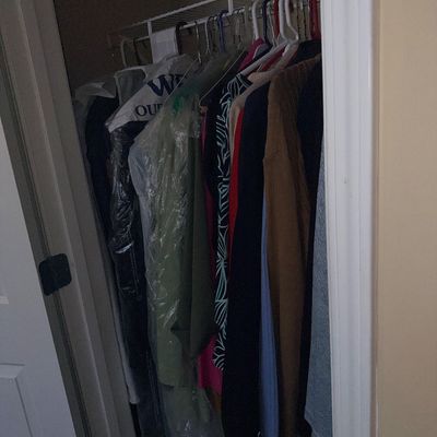 Closet filled with hanging men's and women's clothing, including jackets, sweaters, and dry-cleaned garments on wire racking.