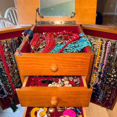 Standing wooden jewelry armoire with mirrored top lid, filled with a large collection of colorful costume necklaces and earrings.