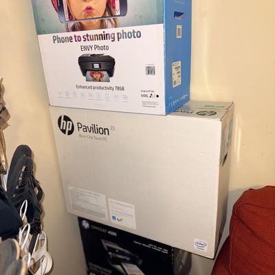 Stack of boxed electronics including an HP Envy Photo 7858 printer and an HP Pavilion 23 All-in-One Touch PC.