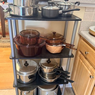 A four-tier black and silver utility shelf displayed with various stainless steel cookware and vintage amber Vision cookware.