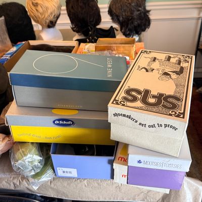 Stack of assorted branded shoe boxes including Nine West, SAS, Dr. Scholl's, and Mootsies Tootsies, displayed alongside several wigs...