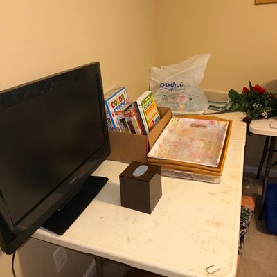 A Sanyo flat-screen television arranged on a white folding table alongside puzzle books, decorative trays, and a tissue box cover.