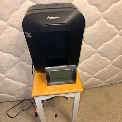 Black Fellowes paper shredder sitting on a small wooden side table alongside a digital weather station display.
