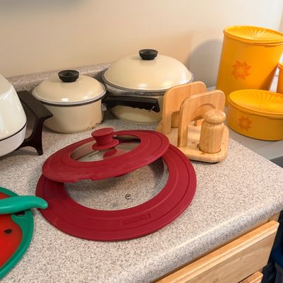 Collection of vintage and modern kitchenware including cream-colored enamel cookware, yellow Tupperware canisters, a wooden napkin...