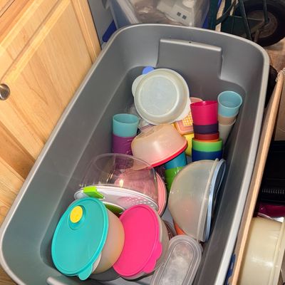 Large gray plastic storage tote filled with various colorful plastic kitchenware, including Tupperware containers, bowls, and stacked cups.
