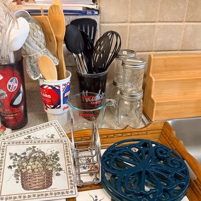 Assortment of kitchenware including a Pyrex glass measuring cup, Mason jars, wooden and plastic utensils, a cast iron trivet, ceramic hot...