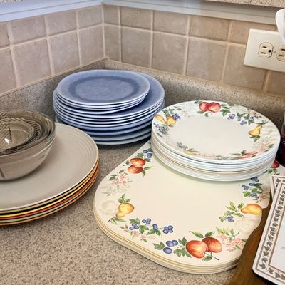 Collection of assorted kitchenware including stacks of blue ceramic plates, fruit-patterned dinnerware with matching placemats, and a set...