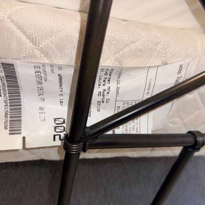 Detail of a mattress tag showing manufacturer information from Ackerman Mfg. Co., positioned behind a black metal bed frame.