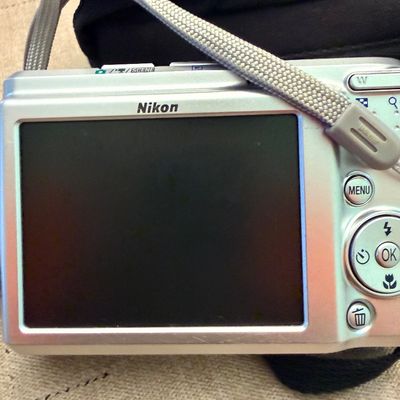 Silver Nikon Coolpix digital point-and-shoot camera featuring a large rear LCD screen and grey wrist strap, resting on a soft case.