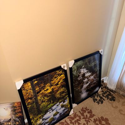 Two framed nature photography prints depicting forest streams, shown leaning against a wall with protective corner guards.