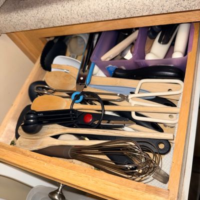 Assorted kitchen utensils in a wooden drawer, including wooden spoons, a metal whisk, tongs, and measuring spoons.