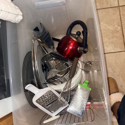 Plastic storage tote filled with assorted modern kitchen utensils, including a red tea kettle, handheld graters, wire whisk, strainers,...