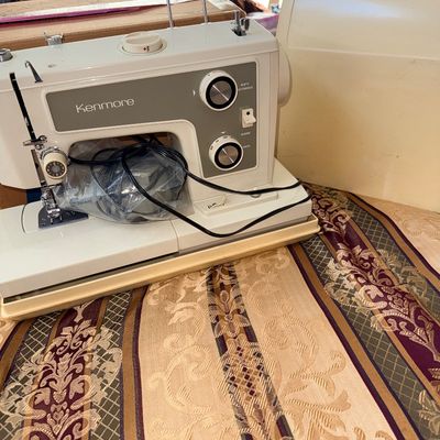 Vintage Kenmore electric sewing machine with off-white body, gray faceplate, power cord, and hard plastic carrying case.