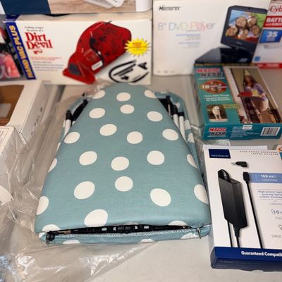 Foldable tabletop ironing board with light blue polka dot cover, shown alongside boxed household electronics and accessories.