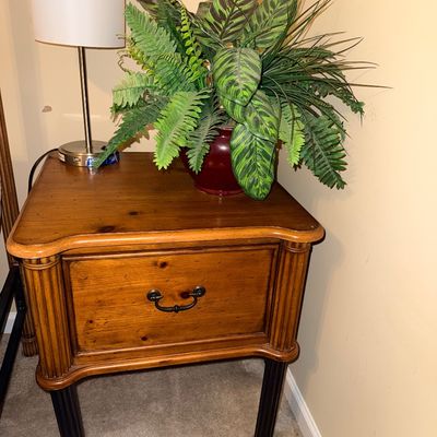 Traditional wooden nightstand featuring a single drawer with a metal pull, fluted corner details, and dark turned legs, topped with a...