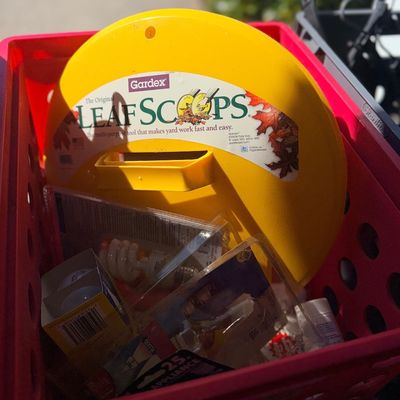 A red plastic crate filled with household items, featuring a bright yellow Gardex Leaf Scoops yard tool alongside packaged light bulbs.