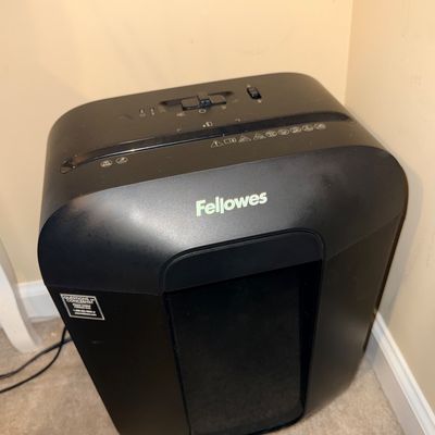 Black Fellowes brand paper shredder sitting on carpeted floor in a corner.