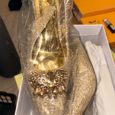 Pair of gold glitter pointed-toe pumps adorned with beaded toe embellishments, wrapped in protective plastic inside a shoe box.