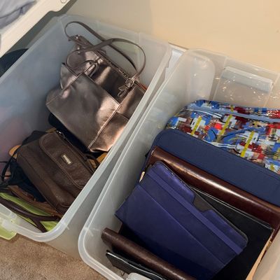 Two plastic storage bins filled with assortment of handbags, purses, and tablet or planner covers.