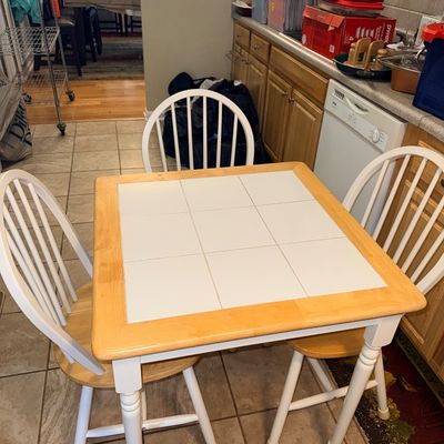 Square light wood dinette table with a white tile insert top, accompanied by three matching white spindle-back dining chairs.