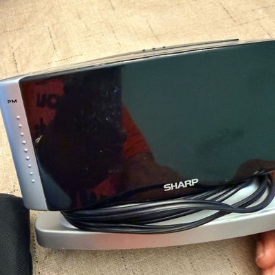 Sharp digital alarm clock with a silver and black casing, shown with attached power cord wrapped around the base.