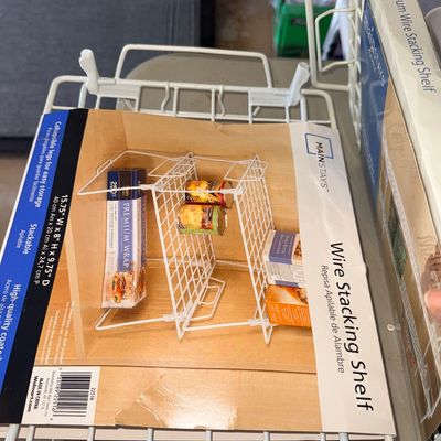 Mainstays white coated wire stacking shelf with original packaging intact, featuring collapsible legs for pantry storage.