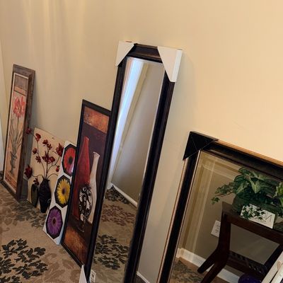 Collection of framed wall art prints and full-length dark-wood framed mirrors leaning against a wall.