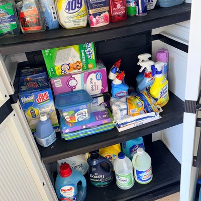 Black plastic storage cabinet stocked with assorted household cleaning supplies, laundry detergents, and floor care products.