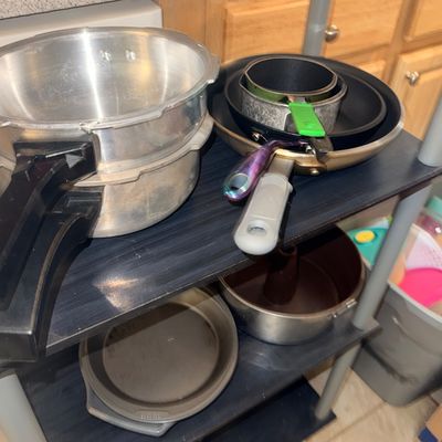 Assorted cookware collection including aluminum pressure cookers, non-stick skillets, small saucepans, pie pans, and a bundt pan arranged...