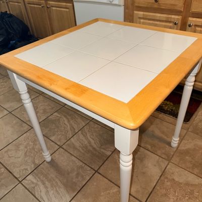 Square white tile-top dining table featuring a light wood frame and white turned legs.