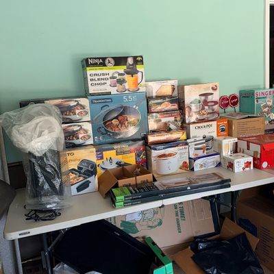 Collection of boxed small kitchen appliances and cookware on folding tables, including a Ninja Master Prep blender, Tramontina 5.5-quart...