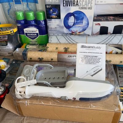 SteamBuddy handheld garment steamer in original plastic packaging with instruction manual, displayed alongside household items and boxed...